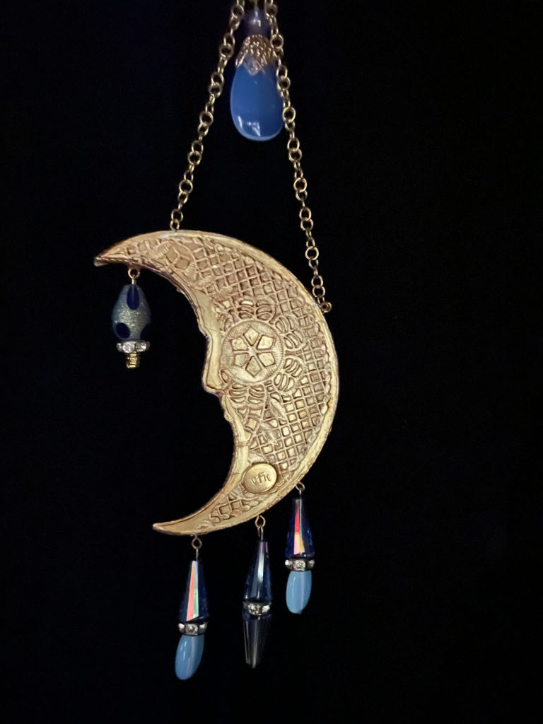 A warm gold tone crescent moon pendant is embellished with Opalite and blue cone-shaped crystals.