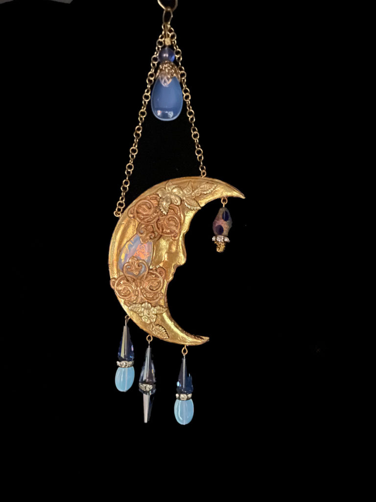 A warm gold tone crescent moon pendant is embellished with Opalite and blue cone-shaped crystals.