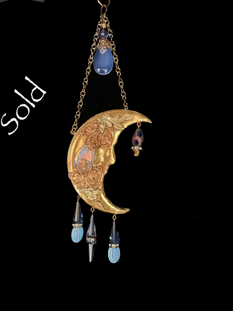 A warm gold tone crescent moon pendant is embellished with Opalite and blue cone-shaped crystals.